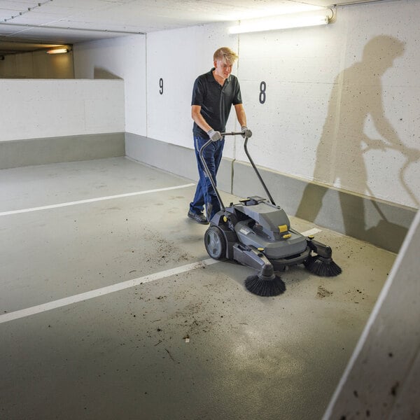 A person is using a large, battery-operated walk-behind floor sweeper to clean debris from the floor of a parking garage.