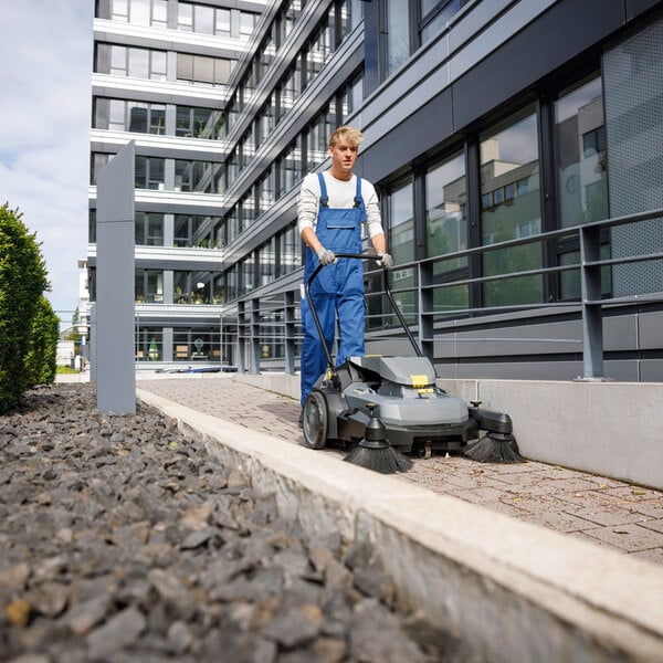 A person in blue overalls is operating a walk-behind floor sweeper on a paved pathway outside a modern building.