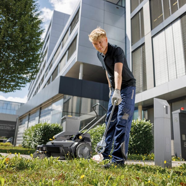 A person is using a grabber tool to pick up trash from the grass in front of a modern building, with a walk-behind floor sweeper nearby.
