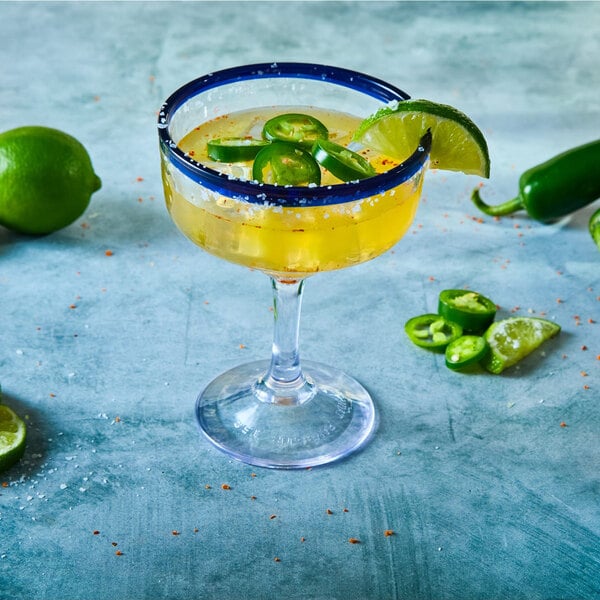 A margarita glass with a blue rim, filled with a yellow drink and garnished with lime and jalapeño slices, surrounded by limes and jalapeños on a blue surface.