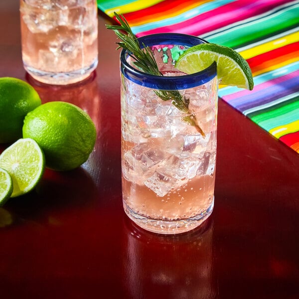 A clear glass with a blue rim filled with a light pink beverage, ice cubes, a lime wedge, and a sprig of rosemary, placed on a red surface next to whole and sliced limes and a colorful striped cloth.