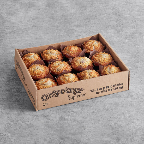 A cardboard box containing twelve blueberry crumb cake muffins arranged in three rows on a gray surface.