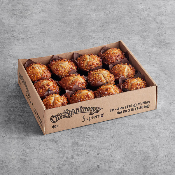 A cardboard box containing twelve golden brown muffins with a crumbly topping, each in a brown paper liner, displayed on a gray surface.