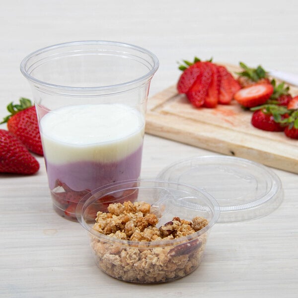 A clear plastic cup filled with layered yogurt and fruit, accompanied by a separate small container of granola, with fresh strawberries and a cutting board in the background.