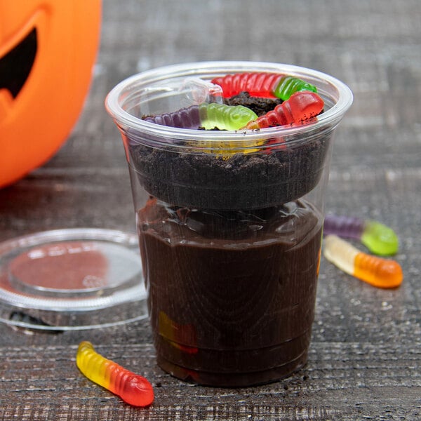 A clear plastic cup filled with chocolate pudding, topped with crushed cookies and colorful gummy worms, with a plastic lid and an orange pumpkin container partially visible in the background.