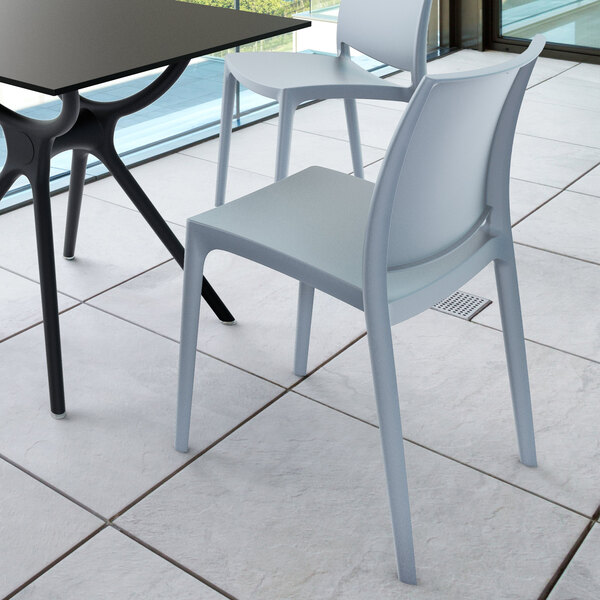 Two modern, light gray chairs with simple, sleek lines are placed next to a black table on a tiled floor near large windows.