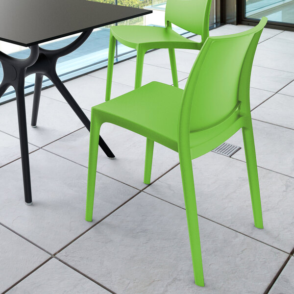 Two bright green plastic chairs are placed next to a black table on a tiled floor in a modern indoor or outdoor setting.