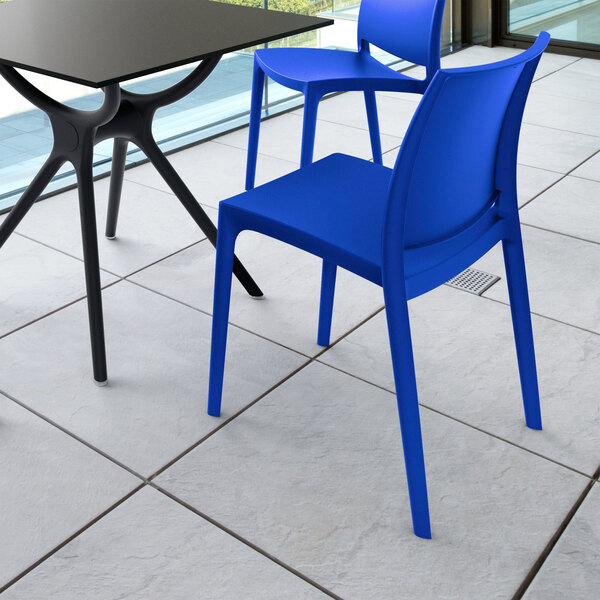 Two modern blue chairs and a black table are placed on a tiled floor near large windows.