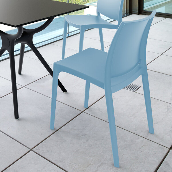 A light blue plastic chair with a simple, modern design is placed next to a black table on a tiled floor near large windows.