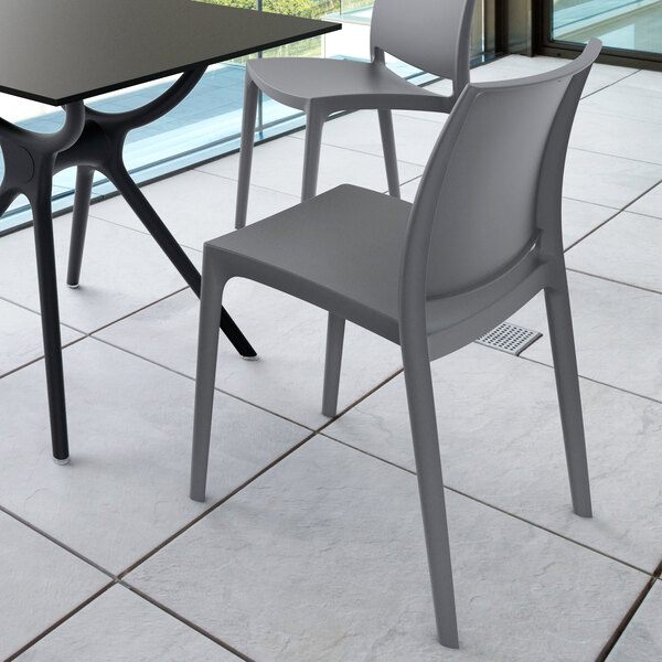 Two modern gray chairs with simple, sleek designs are placed next to a black table on a tiled floor near large windows.