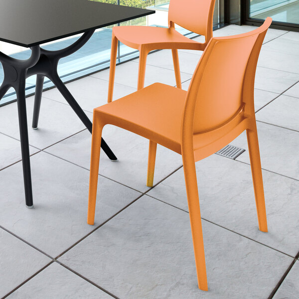 Two orange chairs with a modern design are placed next to a black table on a tiled floor near large windows.