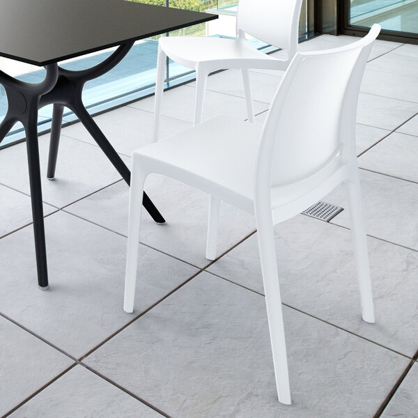 A modern white chair with a curved backrest and four straight legs, placed next to a black table on a tiled floor in a bright indoor space.