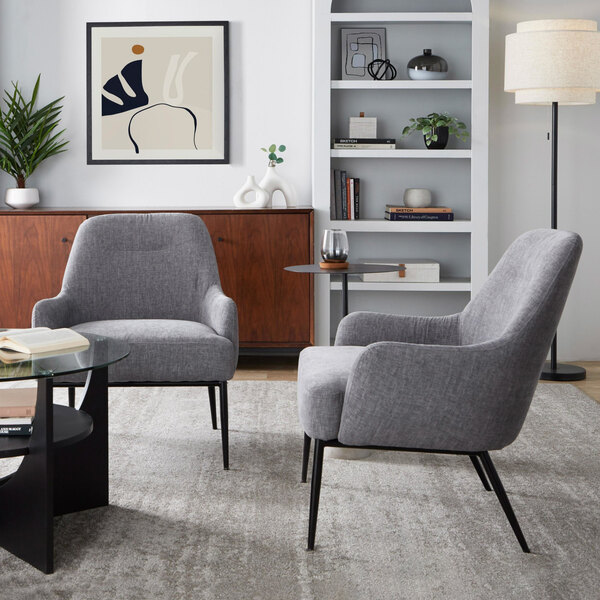 Two modern accent chairs with light grey fabric upholstery and black metal legs are placed in a stylish living room setting with a glass coffee table, a wooden sideboard, a bookshelf, and a floor lamp.