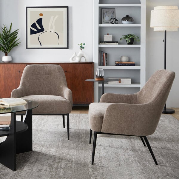 Two light brown fabric accent chairs with black metal legs are placed in a modern living room setting, featuring a glass coffee table, a wooden sideboard, a bookshelf, and a floor lamp.