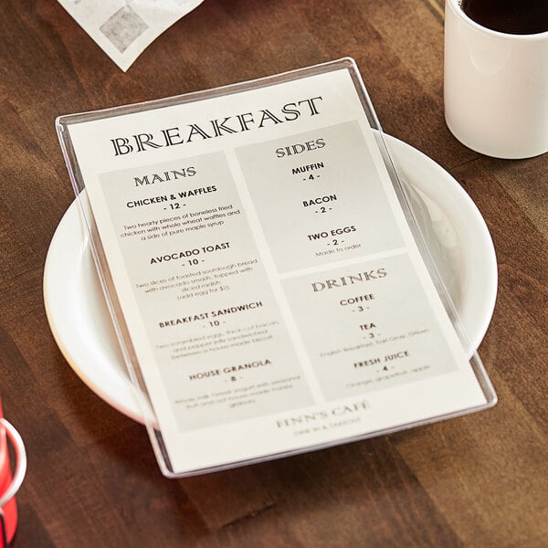 A clear, single-panel menu cover displaying a breakfast menu, placed on a white plate on a wooden table next to a cup of coffee.