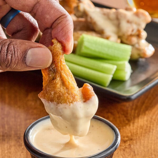 A hand is dipping a chicken wing into a creamy white sauce, with celery sticks and more wings visible in the background.