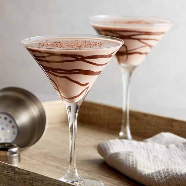 Two martini glasses filled with a creamy, light brown beverage decorated with chocolate swirls, placed on a wooden tray with a cocktail shaker and a white cloth nearby.