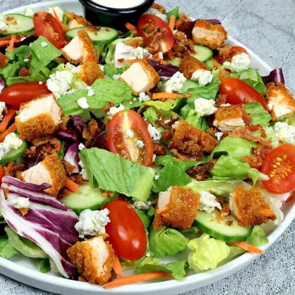 A salad topped with breaded chicken pieces, grape tomatoes, cucumber slices, crumbled cheese, and mixed greens, with a cup of dressing in the background.