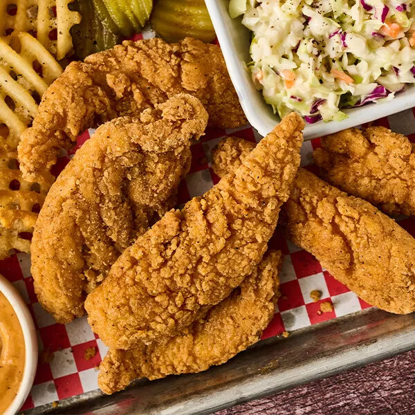 A tray with several pieces of breaded fried chicken tenderloin, waffle fries, pickles, coleslaw, and a dipping sauce.