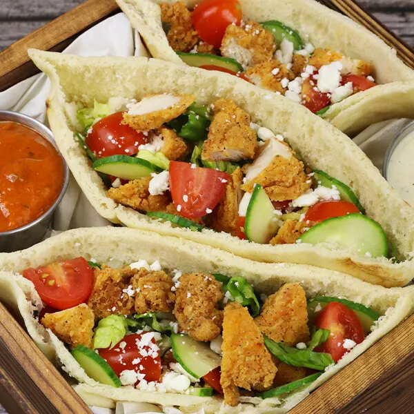 Three pita sandwiches filled with breaded chicken pieces, sliced cucumbers, cherry tomatoes, lettuce, and crumbled cheese, served with two dipping sauces on the side.