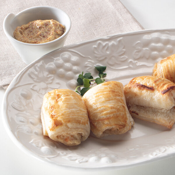 Three golden-brown puff pastry rolls are arranged on a decorative white plate, accompanied by a small sprig of greens, with a cup of grainy mustard dip in the background.