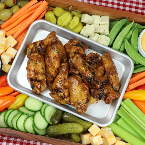 A tray of grilled chicken wings is surrounded by an assortment of fresh vegetables, cheese cubes, and pickles.
