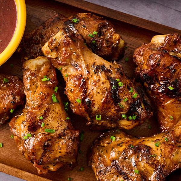 A close-up view of several grilled chicken wings garnished with chopped herbs on a wooden tray, with a small bowl of dipping sauce partially visible.
