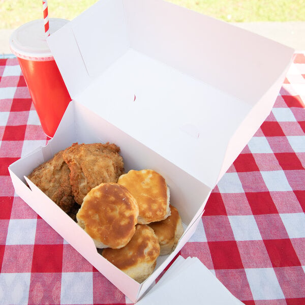 A white takeout box containing fried chicken and biscuits is placed on a red and white checkered tablecloth next to a red cup with a straw.