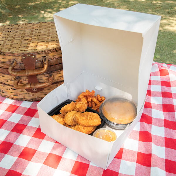 A white takeout box containing fried chicken tenders, crinkle-cut fries, a sandwich in a plastic container, and a dipping sauce, placed on a red and white checkered picnic tablecloth next to a wicker picnic basket.