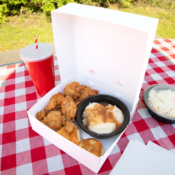 A white takeout box containing fried chicken, mashed potatoes with gravy, and a biscuit is displayed on a red and white checkered tablecloth, accompanied by a drink and a side of coleslaw.