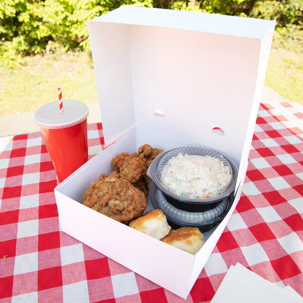 A white takeout box containing fried chicken, biscuits, and a bowl of coleslaw is placed on a red and white checkered tablecloth, with a red drink cup nearby.