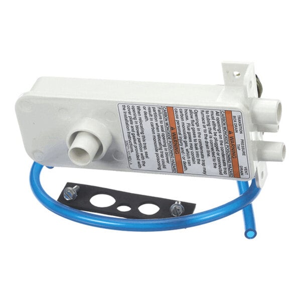 A white plastic component with multiple pipe connections, a blue tube, a mounting bracket, and a warning label.