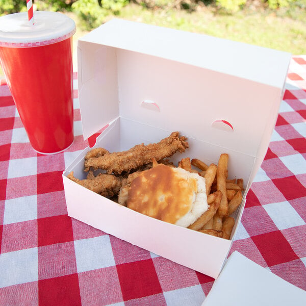 A white takeout box containing fried chicken tenders, French fries, and a biscuit is placed on a red and white checkered tablecloth next to a red cup with a straw.