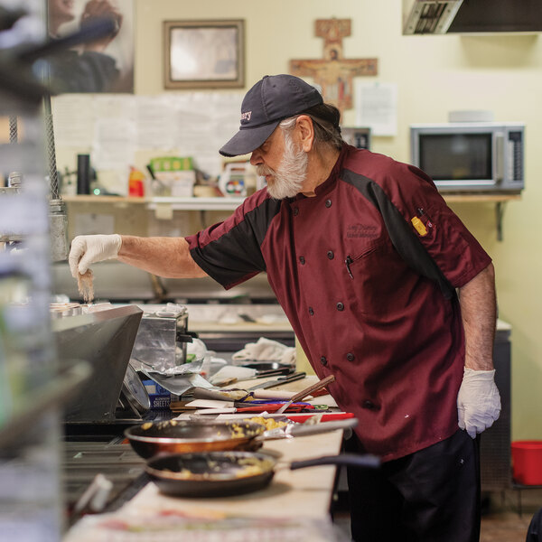 A chef wearing a maroon and black chef coat and a cap is working in a commercial kitchen, seasoning food at a stove.