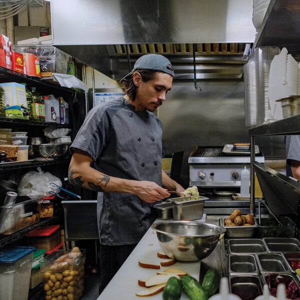 A person wearing a dark short-sleeve chef coat and a backwards cap is preparing food in a commercial kitchen.