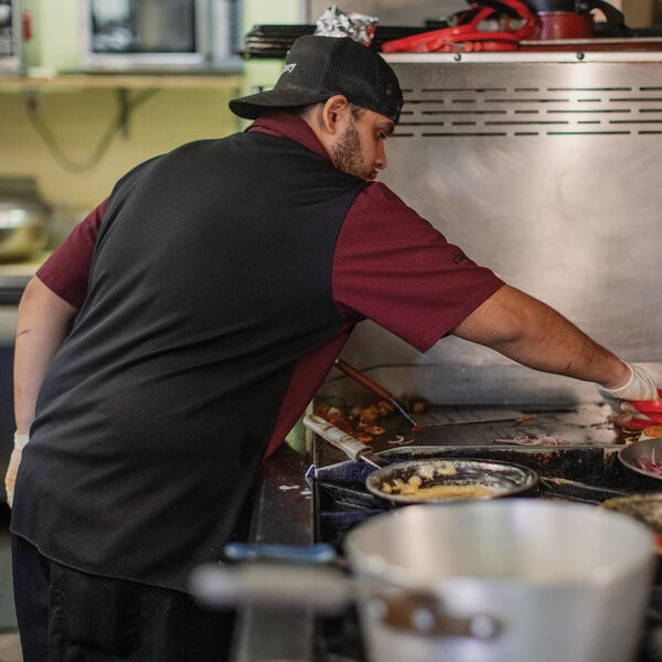 A person wearing a black and maroon short-sleeve shirt and a black cap is cooking at a stovetop in a commercial kitchen.