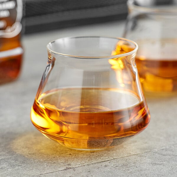A clear, uniquely shaped glass containing an amber liquid, likely whiskey or beer, placed on a gray surface.