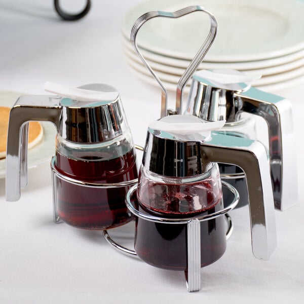 Three glass syrup dispensers with chrome handles are held in a chrome-plated caddy with a heart-shaped handle, placed on a white tablecloth.
