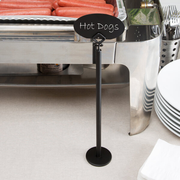 A black metal card holder with an oval sign labeled 'Hot Dogs' stands in front of a buffet tray filled with hot dogs.