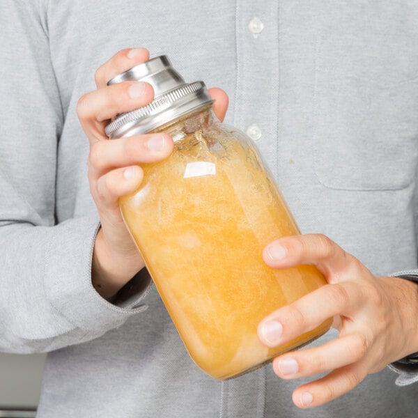 A person is holding and shaking a glass jar filled with a yellowish liquid, fitted with a metal shaker lid.