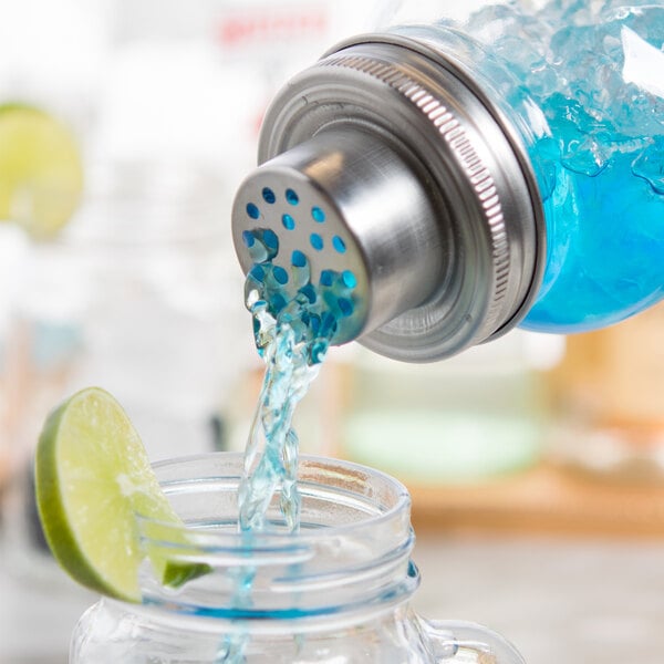 A close-up of a blue liquid being poured from a mason jar cocktail shaker into a glass with a lime wedge.