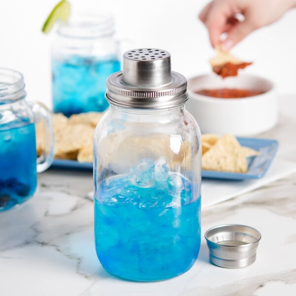 A glass mason jar filled with blue liquid and ice, topped with a metal cocktail shaker lid, sits on a marble surface with snacks and drinks in the background.