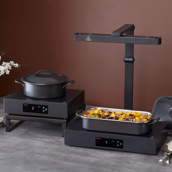 Two black electronic warming trays with digital displays are shown on a countertop, one holding a covered pot and the other holding a rectangular dish of food, with a vertical arm structure above the right tray.