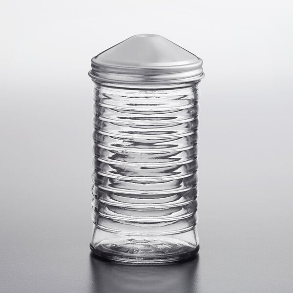 A clear ribbed glass container with a metal lid, designed for pouring, is shown standing upright on a smooth surface.