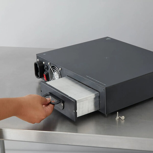 A hand is pulling out a metal drawer from a rectangular black electronic device placed on a stainless steel surface.