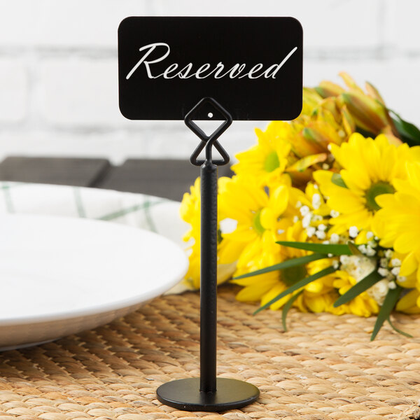 A black metal card holder with a sign that reads 'Reserved' is placed on a woven surface, with a plate and yellow flowers in the background.