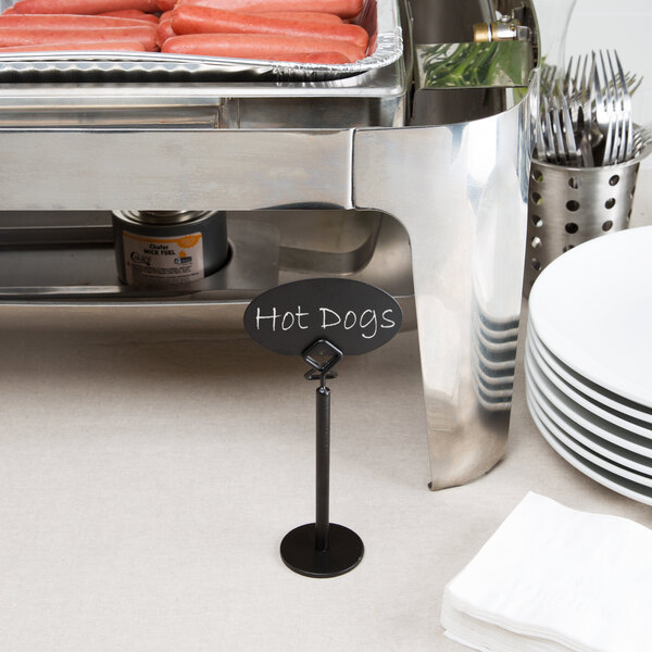 A black metal card holder displaying a sign labeled 'Hot Dogs' is placed on a buffet table next to a chafing dish with hot dogs, a stack of plates, utensils, and napkins.
