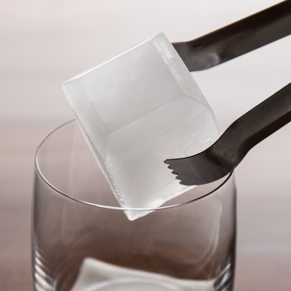 A pair of tongs holding a large, clear ice cube above a glass.