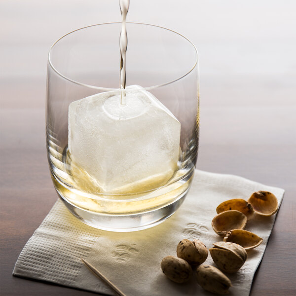 A glass with a large square ice cube being filled with a light-colored liquid, placed on a napkin next to some pistachios and a toothpick.
