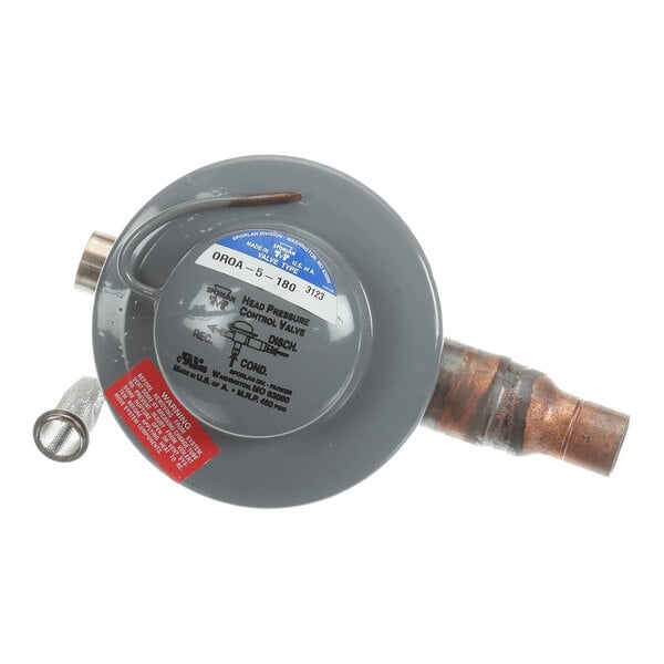 A round, gray metal valve with attached copper piping and labeled stickers, including a blue and white identification label and a red warning label.